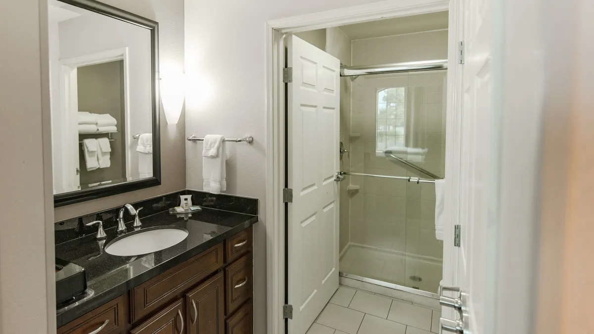 A modern vanity with a spacious sink and a wide mirror sits beside a glass-enclosed shower. The clean, calming design evokes a sense of comfort and the rituals of preparing for or winding down from a busy day.