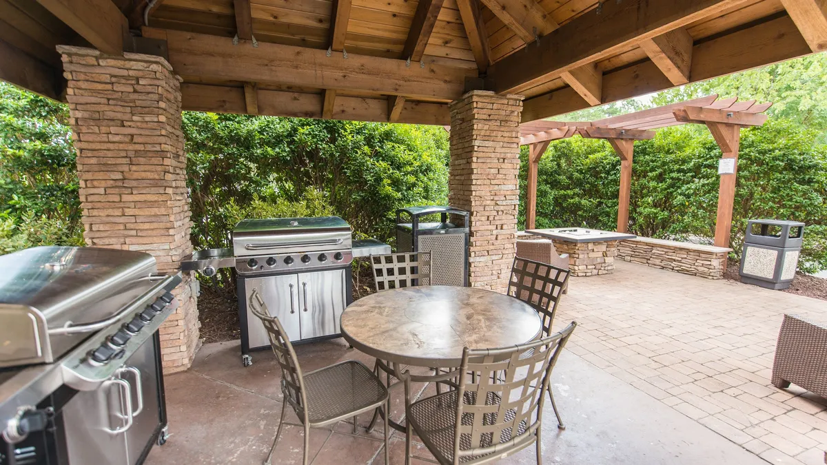 An outdoor grill station complete with a dining table, surrounded by lush greenery and stone columns. The setup encourages outdoor cooking, dining, and social gatherings, highlighting the communal BBQ experience mentioned in the section.