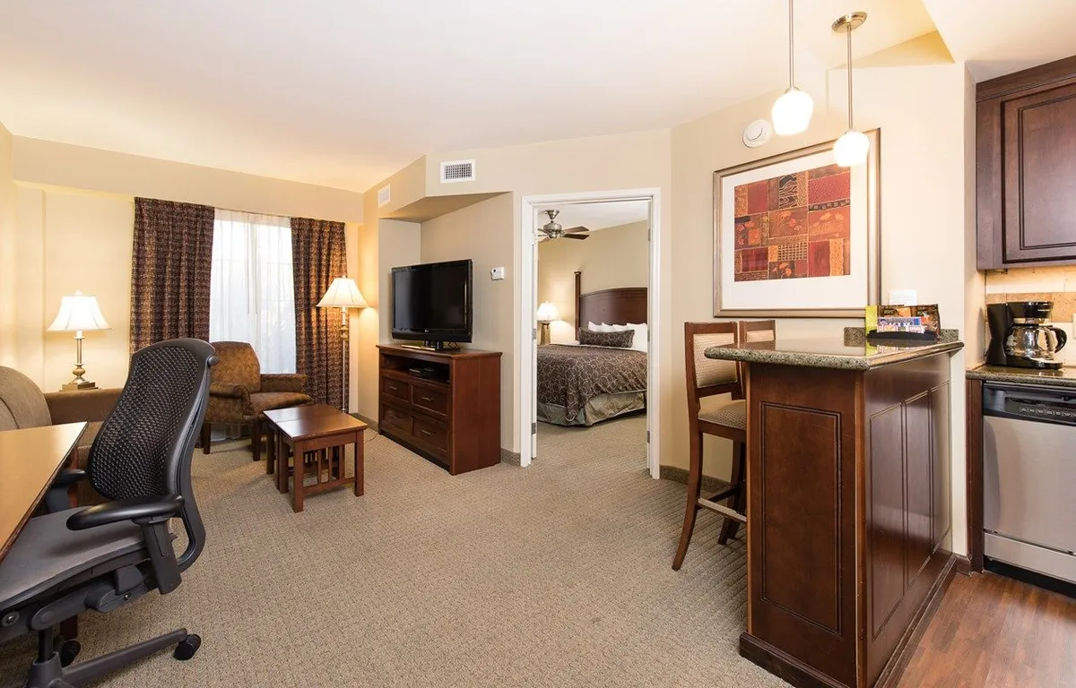 The image showcases a bright, modern condo interior featuring a cozy living area with a sofa and TV, a functional desk for remote work, a well-equipped kitchen area with a bar, and a glimpse into the inviting bedroom. The space is styled for comfort and practicality, ideal for relaxing, working, enjoying meals, or unwinding with loved ones.